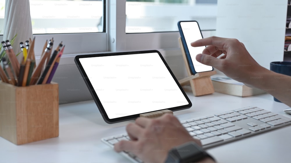 Vista de cerca de un joven que trabaja en línea con una tableta digital en una mesa blanca y usa un teléfono móvil en la oficina en casa.
