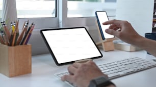 Vista de cerca de un joven que trabaja en línea con una tableta digital en una mesa blanca y usa un teléfono móvil en la oficina en casa.