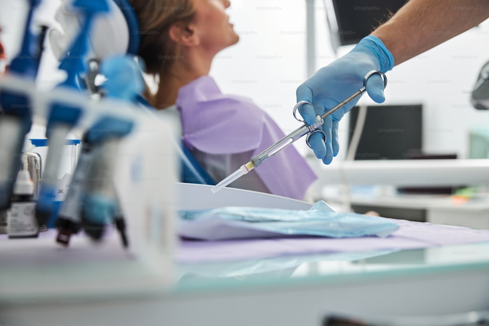 Medical person taking a dental syringe with anesthetic from a table with female patient in a background