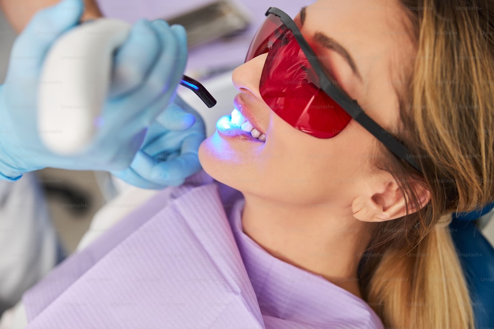 赤いプラスチックの眼鏡で目を覆い、LED硬化ライトの歯科治療を受けながら微笑む女性