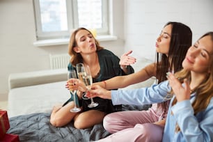 Attractive birthday girl and her female friends with glasses of sparkling wine sitting on the bedspread