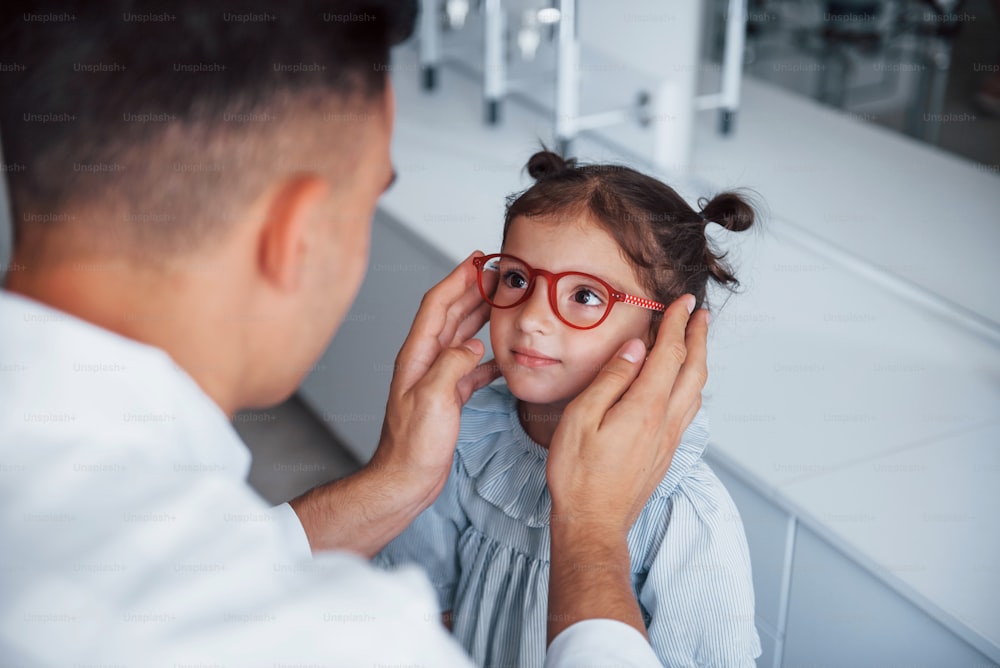 Junge Kinderärztin im weißen Kittel hilft, eine neue Brille für ein kleines Mädchen zu bekommen.