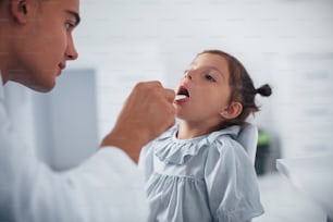 Uses tonsil to check throat. Young pediatrician works with little female visitor in the clinic.