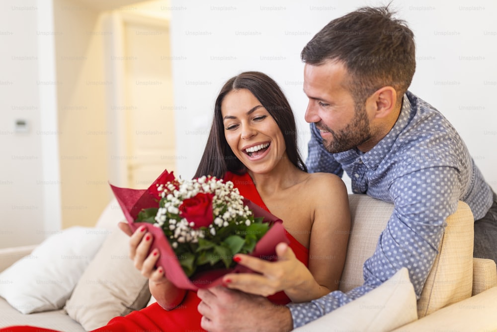 Excited young woman receiving unexpected Bouquet of red roses from husband at home, loving generous boyfriend making romantic surprise to attractive girlfriend on Valentines day occasion