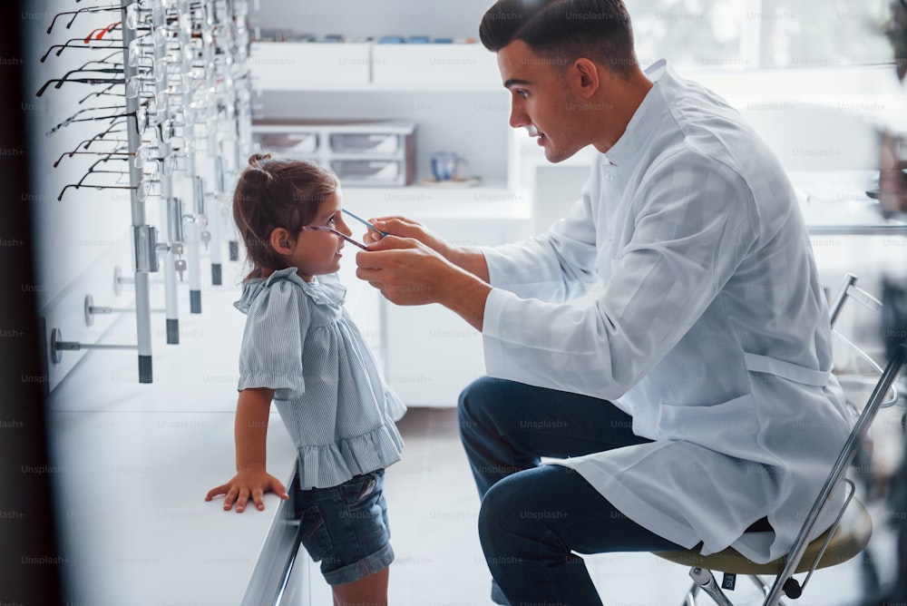 Young pediatrician in white coat helps to get new glasses for little girl.