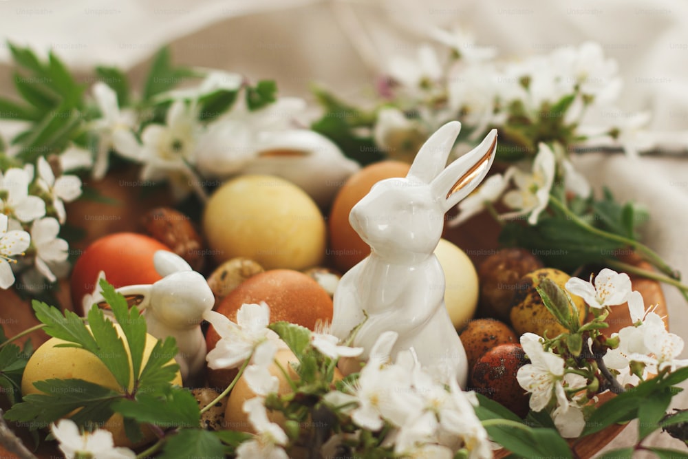 ¡Felices Pascuas! Huevos de pascua modernos con flores de primavera y conejito en cuenco de madera sobre tela de lino rústico. Huevos teñidos al natural sobre textil gris con flores en flor, anémona y liebre. Estético