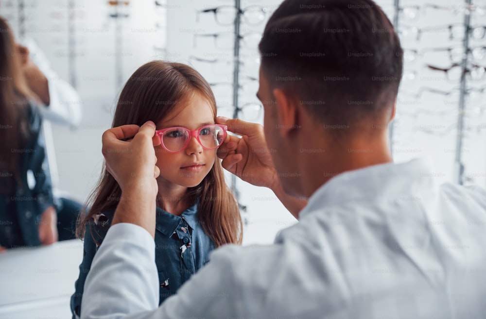 Young pediatrician in white coat helps to get new glasses for little girl.