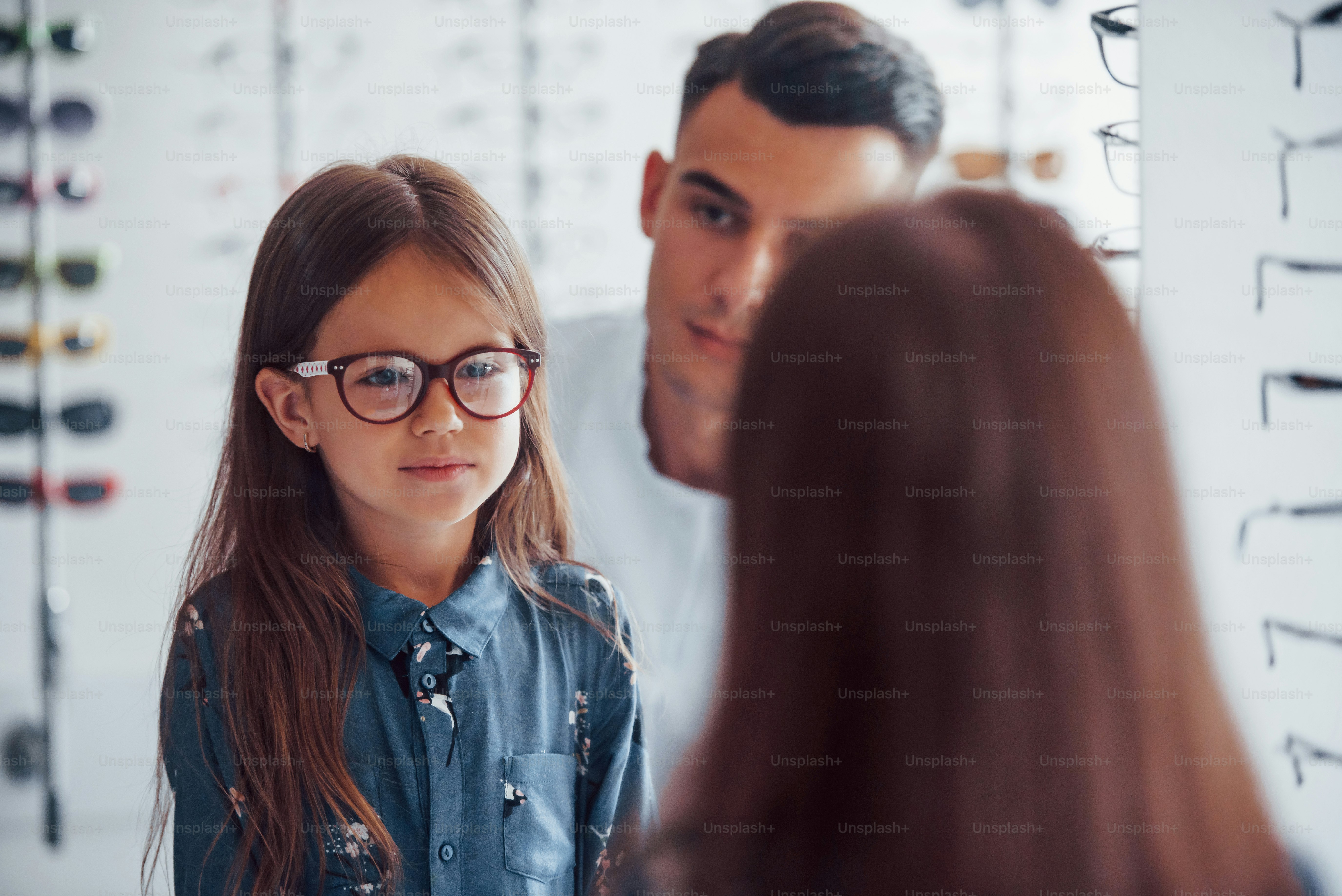 Young pediatrician in white coat helps to get new glasses for little girl.