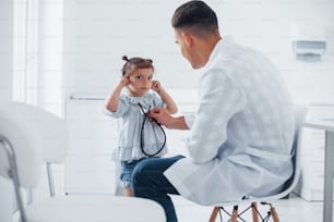 Teaches girl how to use stethoscope. Young pediatrician works with little female visitor in the clinic.
