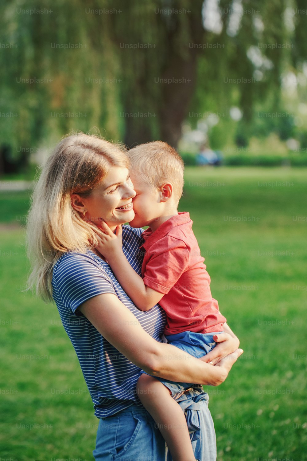 Young smiling Caucasian mother holding cuddling with boy toddler son. Laughing mom hugging embracing child outdoor in park on summer day. Happy authentic family childhood lifestyle.