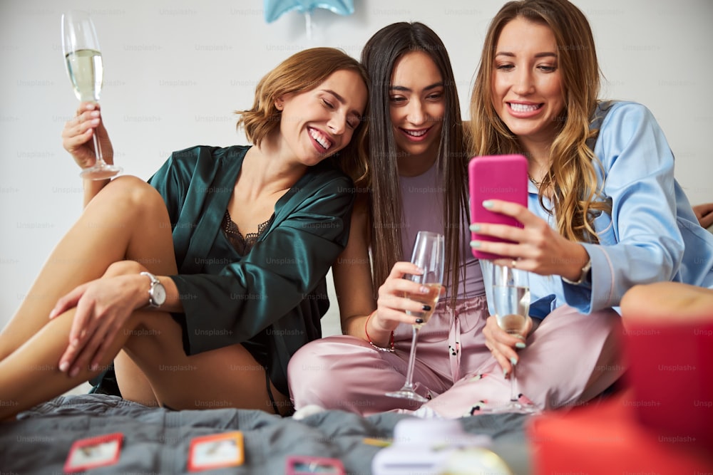 Frontansicht von lächelnden Damen mit Champagnerflöten, die durch die Selfies auf dem Handy schauen