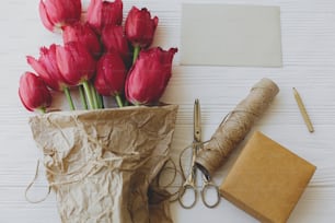 Beautiful red tulips in craft paper with greeting card, gift, pencil and scissors on white wooden background. Rustic  Greeting card template. Happy Mothers day. Happy Women's day.