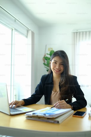 Portrait of cheerful businesswoman in suit working with computer at office desk and smiling to camera.