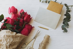 Bonne fête des mères. Belles tulipes rouges en papier kraft avec carte de vœux, cadeau, crayon, ciseaux et ficelle sur fond en bois blanc rustique. Joyeuse Journée de la Femme. Modèle de carte de vœux