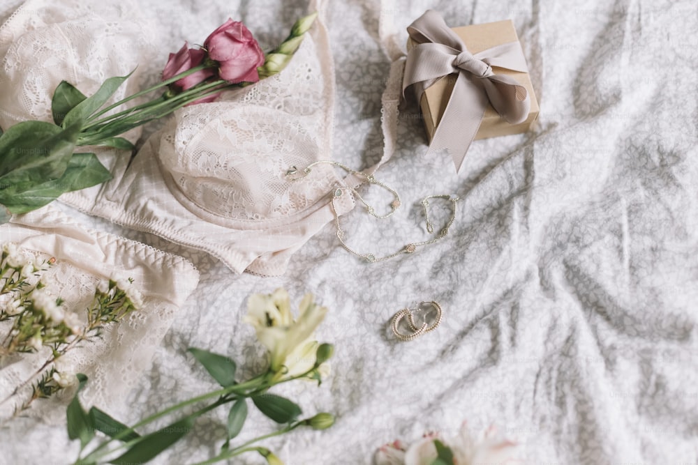 Stylish lace lingerie, gift box, modern jewelry and spring bouquet  on bed. Soft tender feminine image, sensual mood at home concept. Happy Women's day, woman essentials