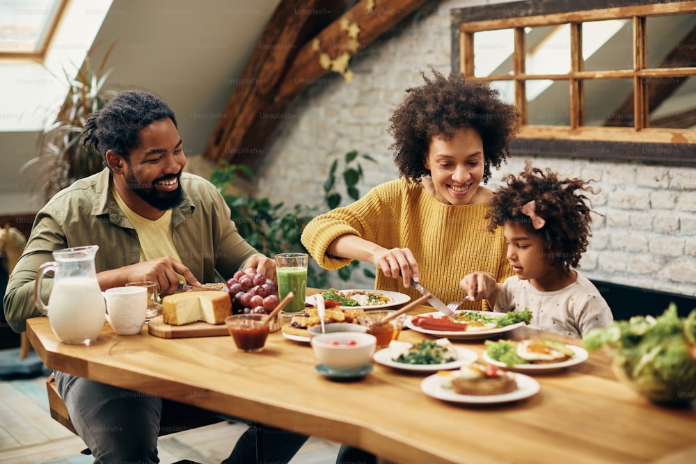 Padres negros felices y su hija comiendo en la mesa del comedor de su casa.