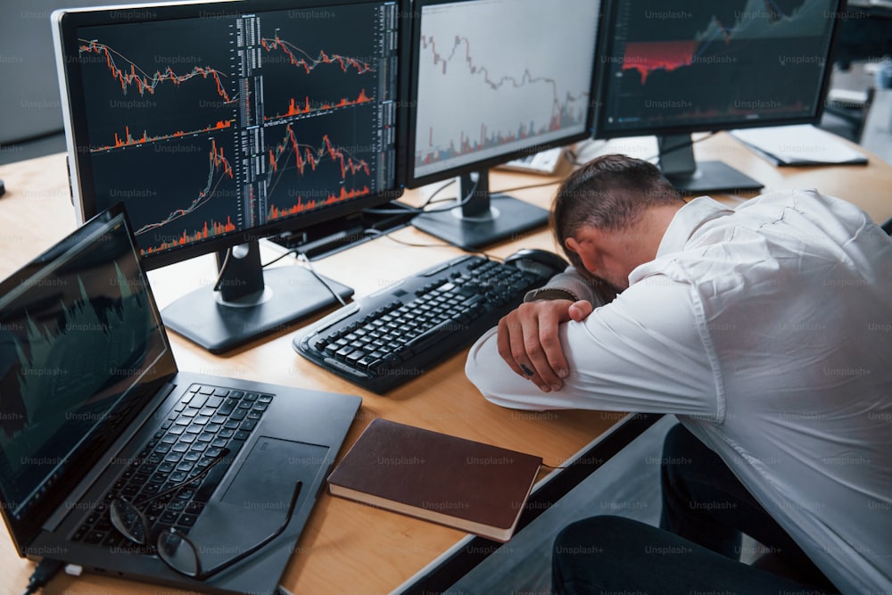 Tired businessman sleeping on the workplace by leaning on the table with multiple screens on it. Stock information on displays.
