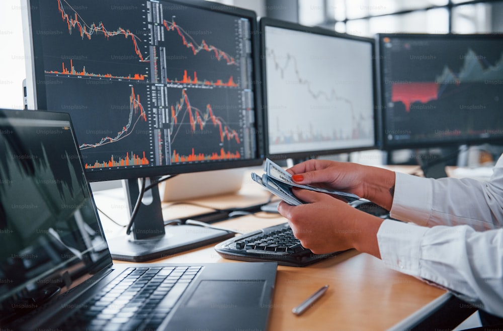 Close up view of woman's hands that holds money near the monitors with graphs.