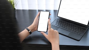 Close up view of young woman holding mobile phone with empty screen while sitting in front of computer tablet at her workplace.