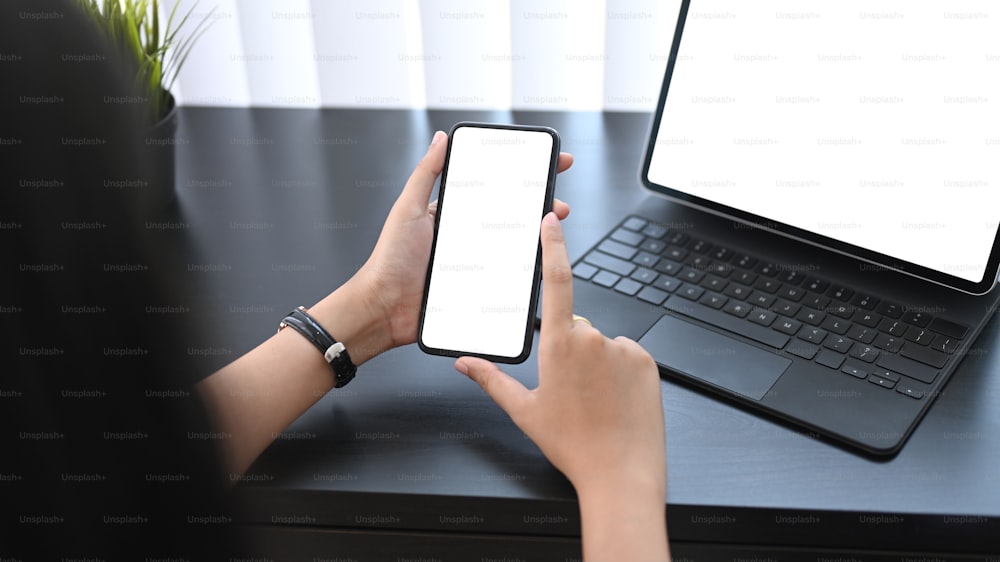 Vue rapprochée d’une jeune femme tenant un téléphone portable avec un écran vide alors qu’elle est assise devant une tablette d’ordinateur sur son lieu de travail.