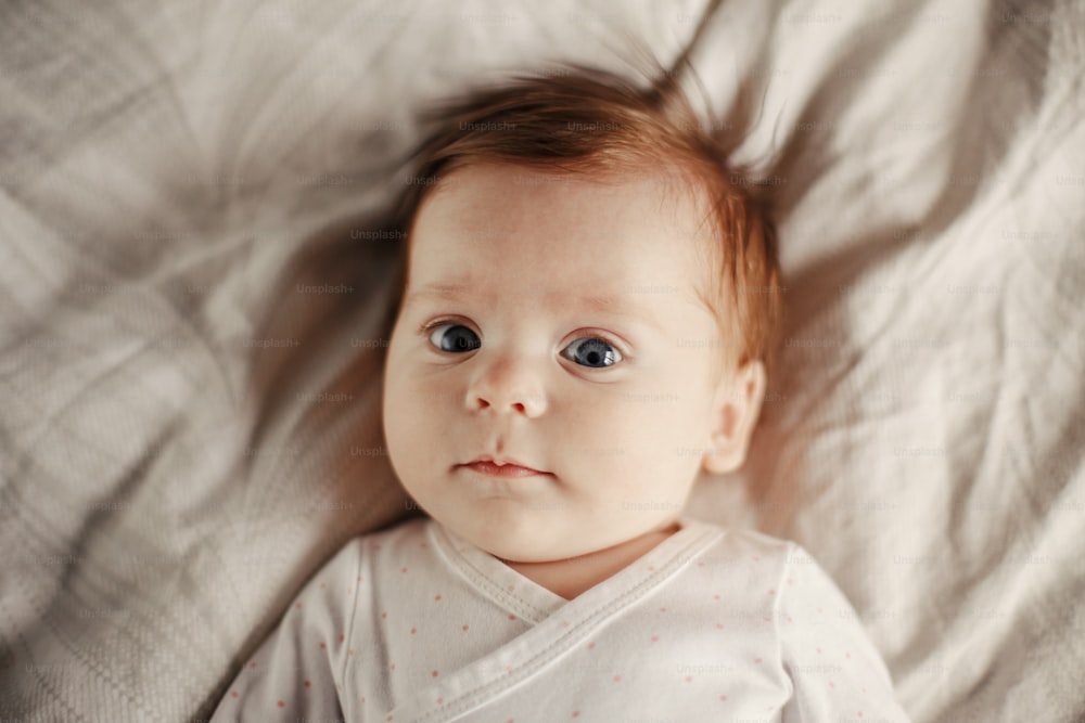 かわいい白人の生まれたばかりの赤ちゃんの接写。青い灰色の目と赤い髪を持つ愛らしい面白い子供の乳児は、カメラを見ながらベッドに横たわっています。本物の子供時代とライフスタイルの率直な瞬間。