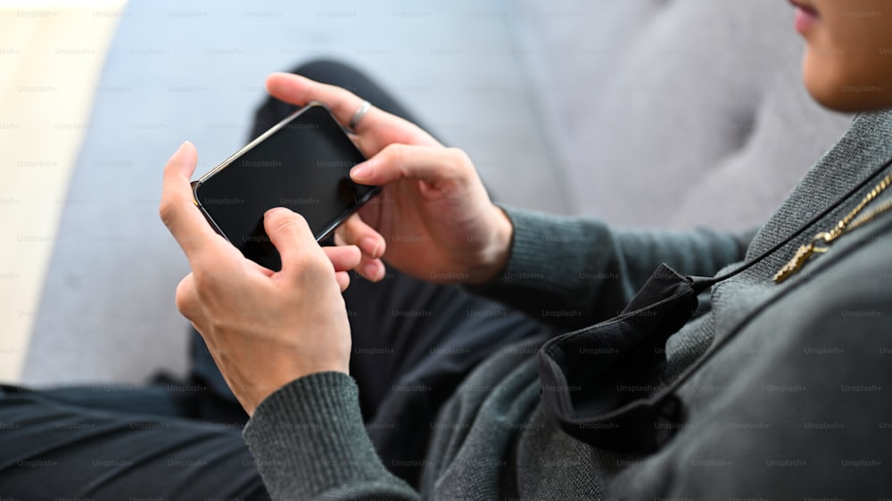 Young man relaxing on comfortable sofa and holding horizontal mobile phone with blank screen.