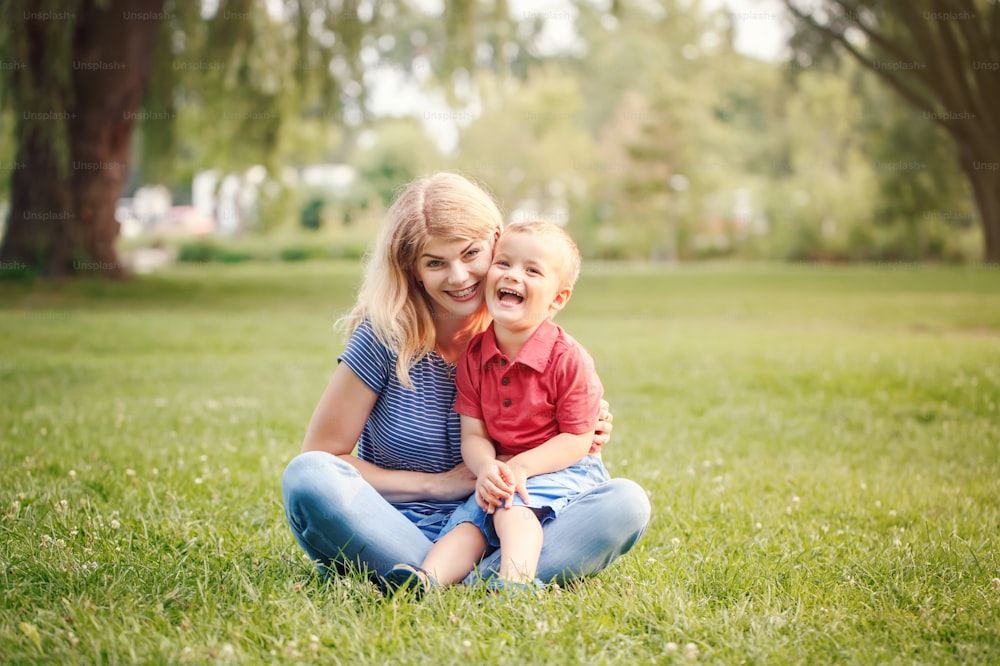 Young smiling Caucasian mother and laughing boy toddler son sitting on grass in park. Family mom and child hugging having fun outdoor on summer day. Happy authentic family childhood lifestyle.
