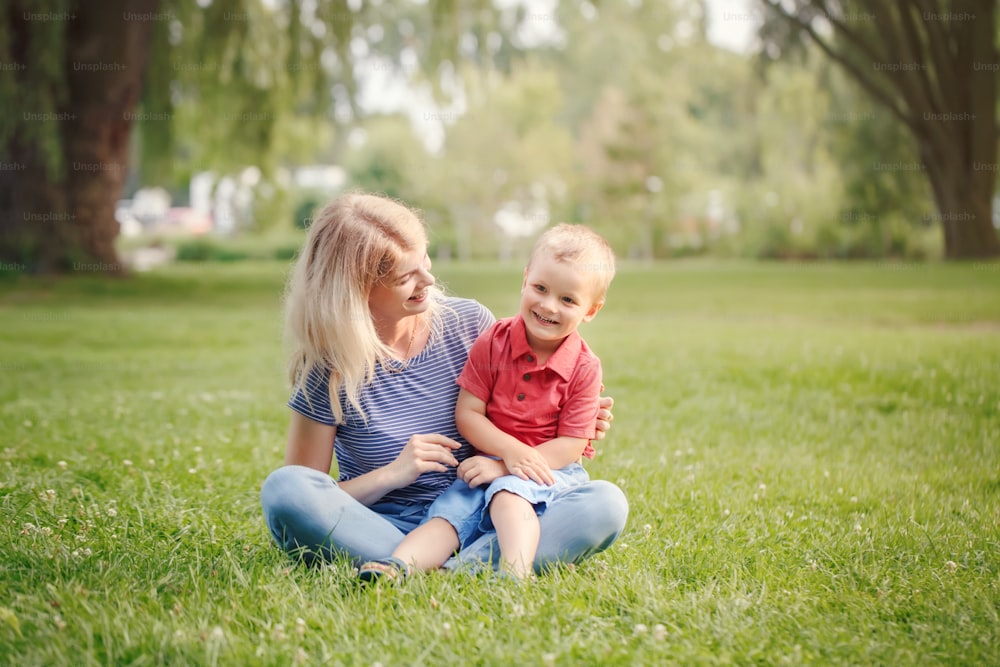 젊은 웃는 백인 어머니와 웃는 소년 유아 아들은 공원의 잔디에 앉아 있습니다. 가족 엄마와 아이는 여름날 야외에서 즐거운 시간을 보내고 있습니다. 행복 한 정통 가족 어린 시절 생활 방식.
