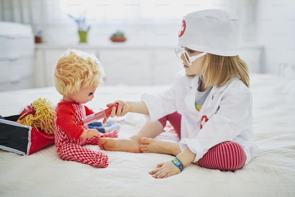 Adorable toddler girl in white coat with syringe playing doctor and giving medical care to her doll. Children and role games. Covid-19 vaccination concept