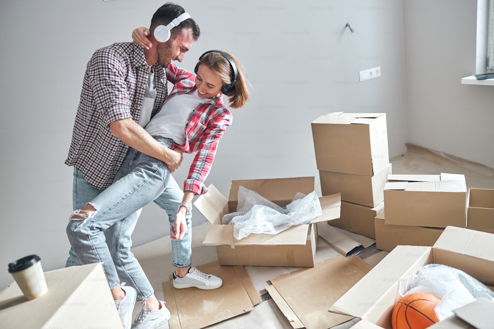Pleased handsome romantic man and his wife in wireless headphones dancing tango among cardboard boxes