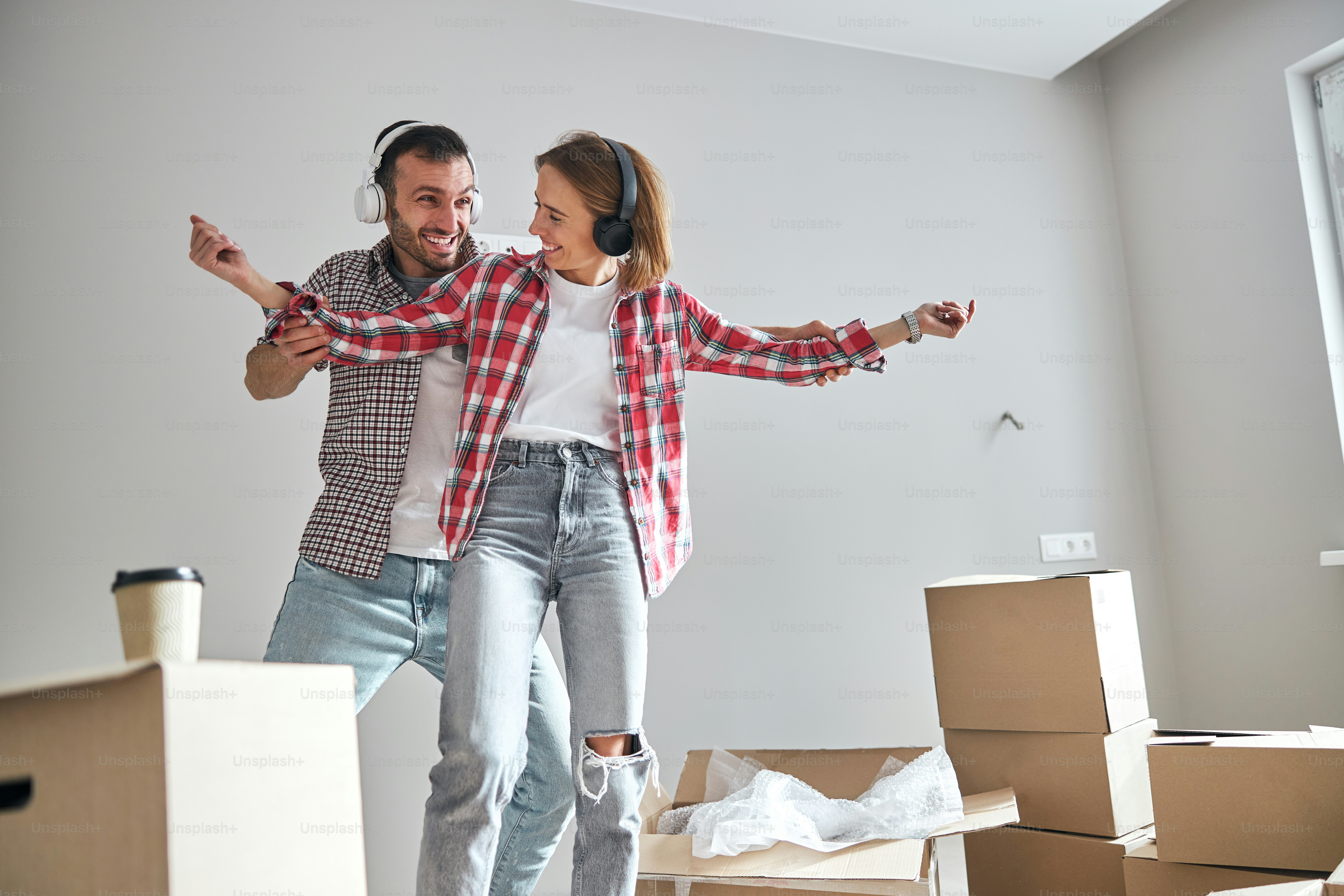 Un homme espiègle et une joyeuse dame dans des écouteurs sans fil s’amusent dans leur nouvel appartement