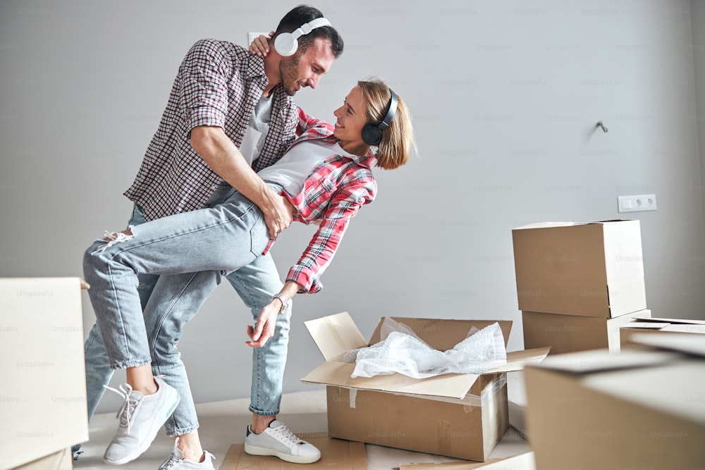 Jovens cônjuges românticos sorridentes em fones de ouvido sem fio realizando uma dança de parceiro em seu novo apartamento