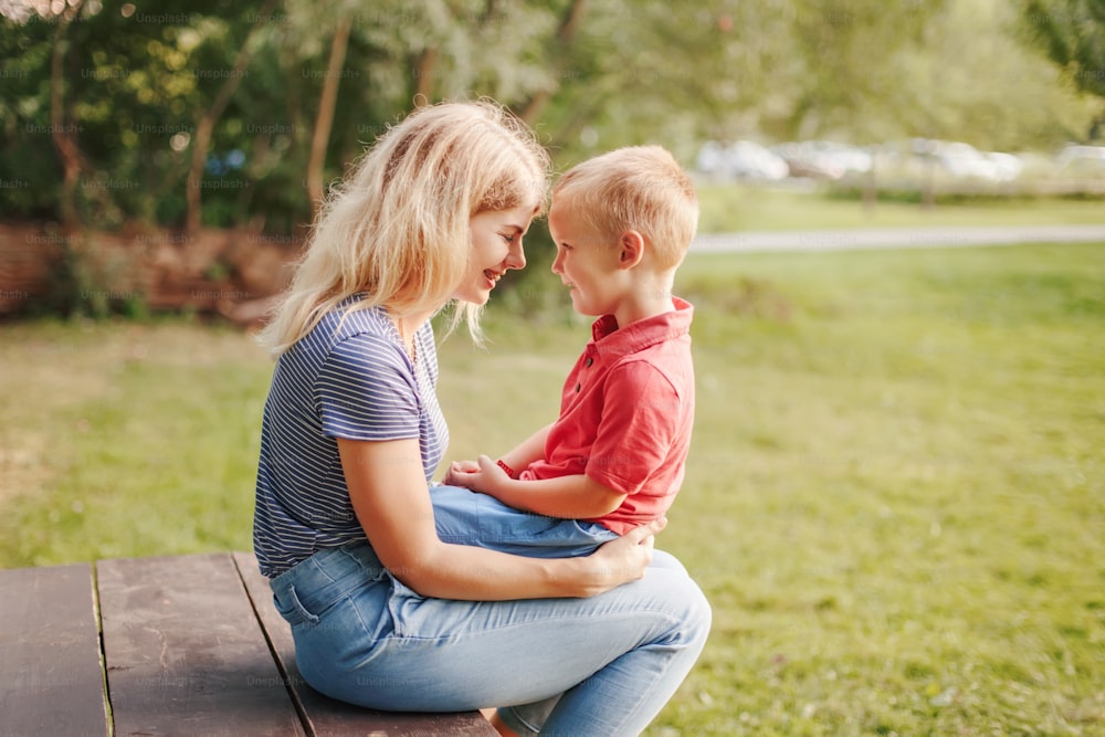 Young Caucasian mother and boy toddler son sitting together face to face. Family mom and child talking communicating outdoor on summer day. Happy authentic family childhood lifestyle.