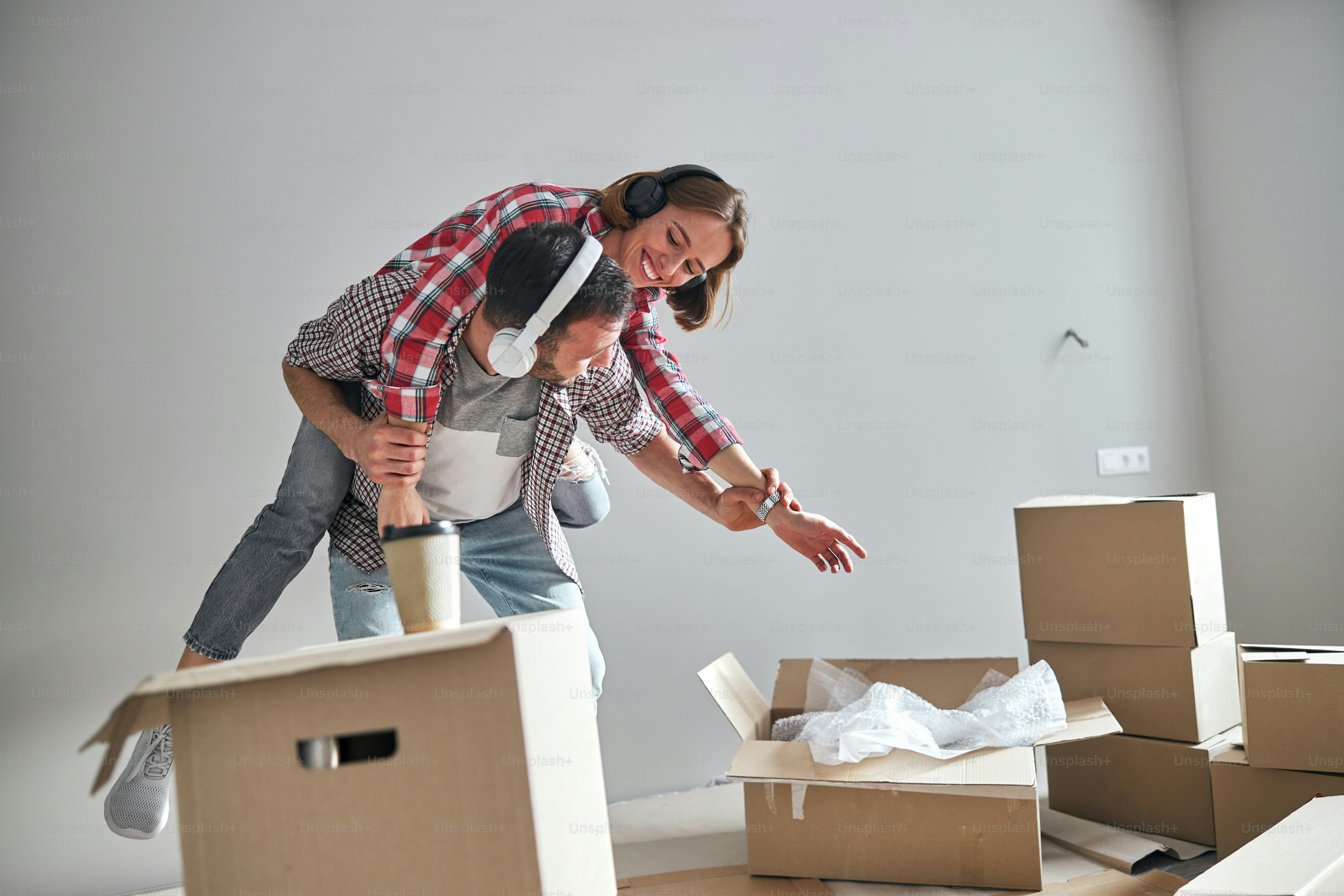 Homme caucasien aux cheveux noirs portant des écouteurs sans fil portant sa conjointe souriante sur son dos dans leur nouvel appartement