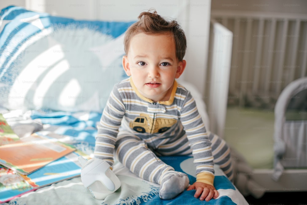 Cute adorable Caucasian kid boy sitting on a bed drinking milk from kids bottle. Healthy eating drinking for little children. Supplementary food for growing babies. Candid real authentic moment.