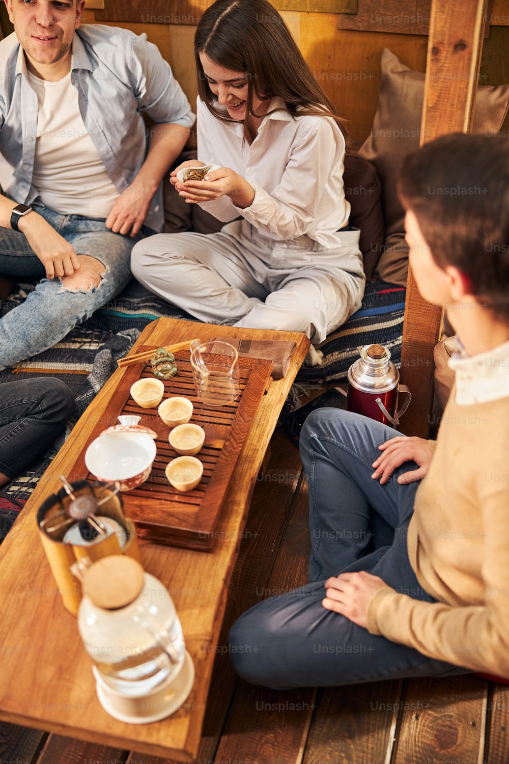 Belle jeune femme tenant un bol de feuilles de thé et souriant tout en étant assise à la table avec des amis dans un café confortable