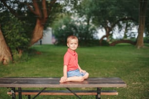 Portrait of cute adorable toddler boy sitting alone in park. Baby child kid sitting outdoor on summer day looking in camera. Pensive kid thinking. Happy authentic childhood lifestyle.