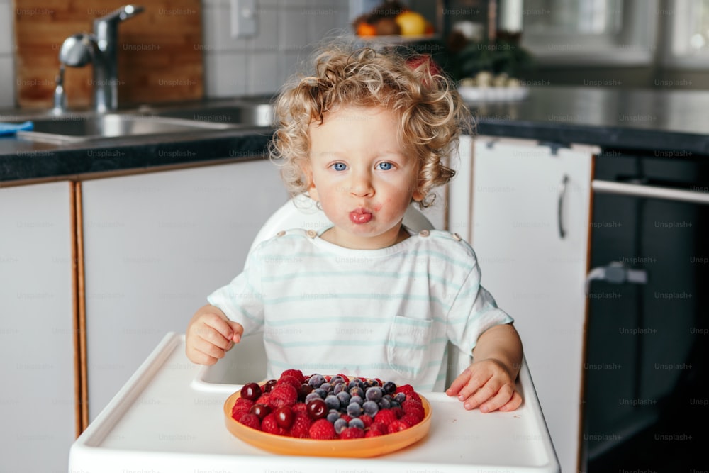 Süßer kaukasischer Junge, der zu Hause reife rote Früchte isst. Lustiges Kind Kind sitzt Hochstuhl mit frischen Beeren in der Küche. Ergänzendes gesundes Fingerfood für Kleinkinder.