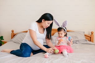 Happy Easter. Asian Chinese pregnant mother with baby girl playing with colorful Easter eggs on bed at home. Kid child and parent celebrating traditional Christian holiday.