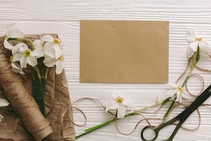 母の日おめでとう。美しい水仙、グリーティングカード、はさみ、白い素朴な木製の背景に麻ひも、テキスト用のスペースとフラットレイ。 女性の日おめでとう。 花のグリーティングカードテンプレート