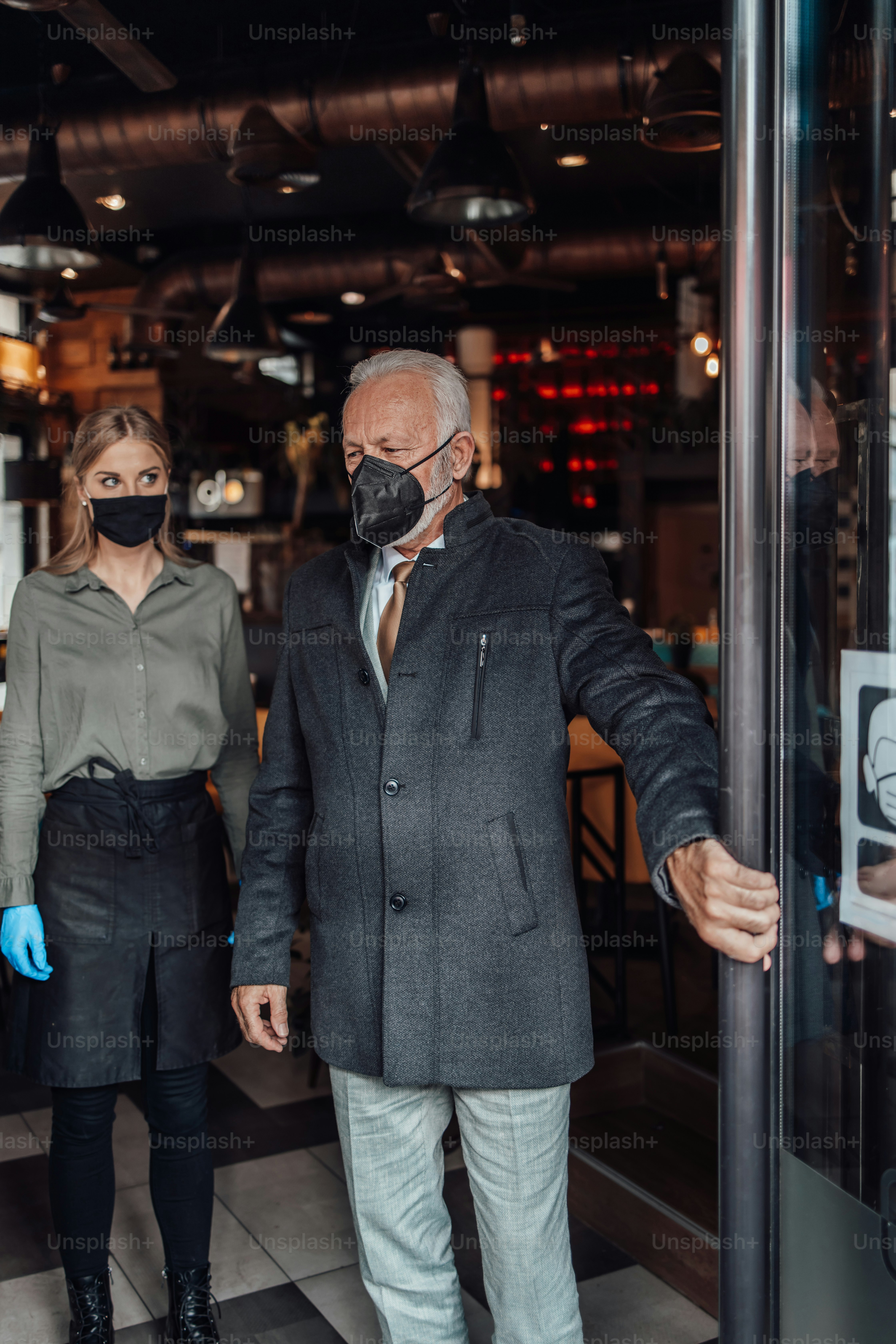 Senior businessman walks out from a restaurant. Young waitress kindly sees him off. They are wearing protective face mask as protection against virus pandemic.