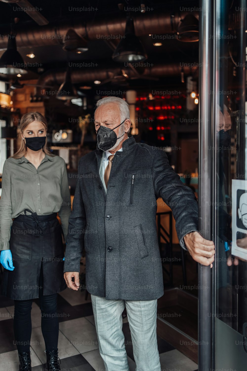 レストランから出てくる年配のビジネスマン。若いウェイトレスが優しく見送ってくれる。彼らはウイルスのパンデミックから身を守るために保護フェイスマスクを着用しています。