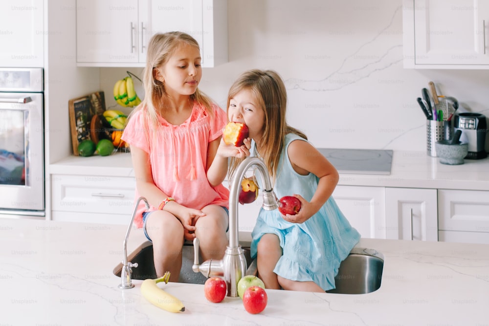Caucasian children girls eating sharing fresh fruits sitting in kitchen sink. Happy family sisters siblings having snack. Organic food healthy delicious tasty meal for kids. Lifestyle authentic moment