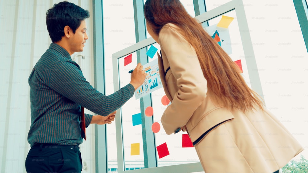 Business people work on project planning board in office and having conversation with coworker friend to analyze project development . They use sticky notes posted on glass wall to make it organized .