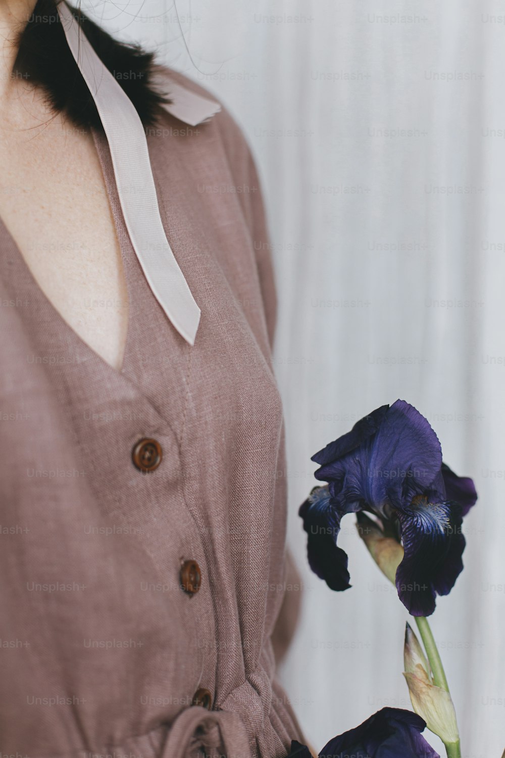 Stylish woman in linen dress holding purple iris flower in sunny light, detail close up. Slow life. Young female in boho rustic dress with iris. Simple aesthetic moment