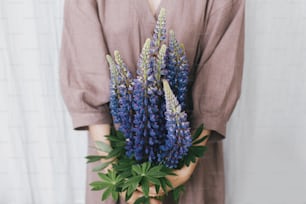 Stylish woman in linen dress holding lupine bouquet in hands closeup at pastel fabric. Simple slow living. Young female in boho rustic dress  with lupine wildflowers. Aesthetics