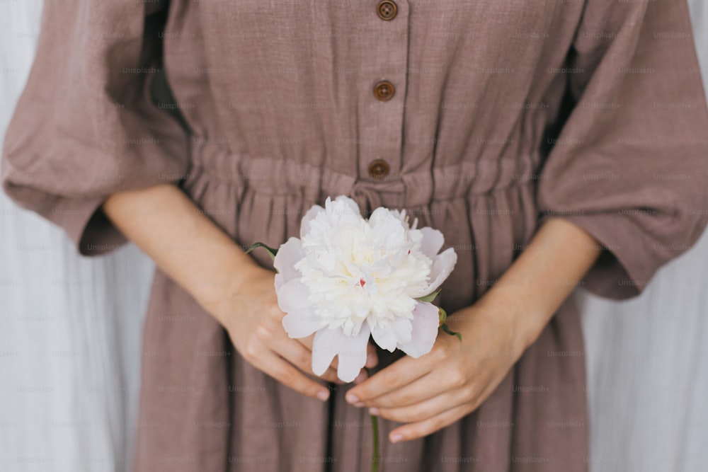 Stylish woman in linen dress holding peony in hands closeup. Slow life. Young female in boho rustic dress with white peony flower. Simple aesthetic moment