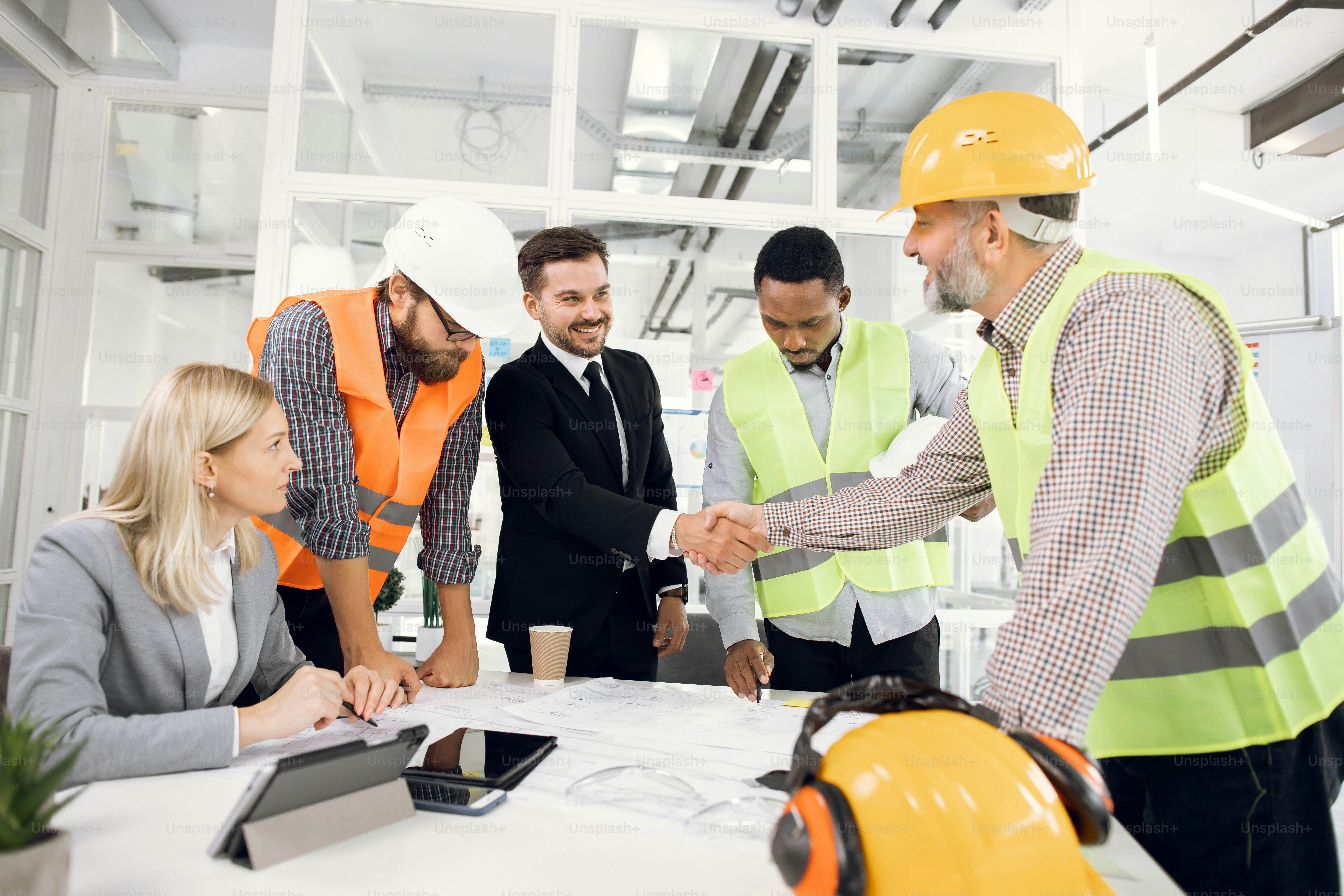 Happy construction team shaking hands during conference at office. Multiracial partners in suits and helmets working with blueprints at office.