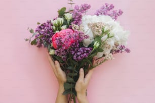 밝은 분홍색 배경에 현대적인 신선한 꽃다발을 들고 있는 손. 세련 된 화려한 인사말 카드에는 모란, 라일락, 유칼립투스, 수국 꽃이 있습니다. 행복한 여성의 날. 해피 어머니의 날