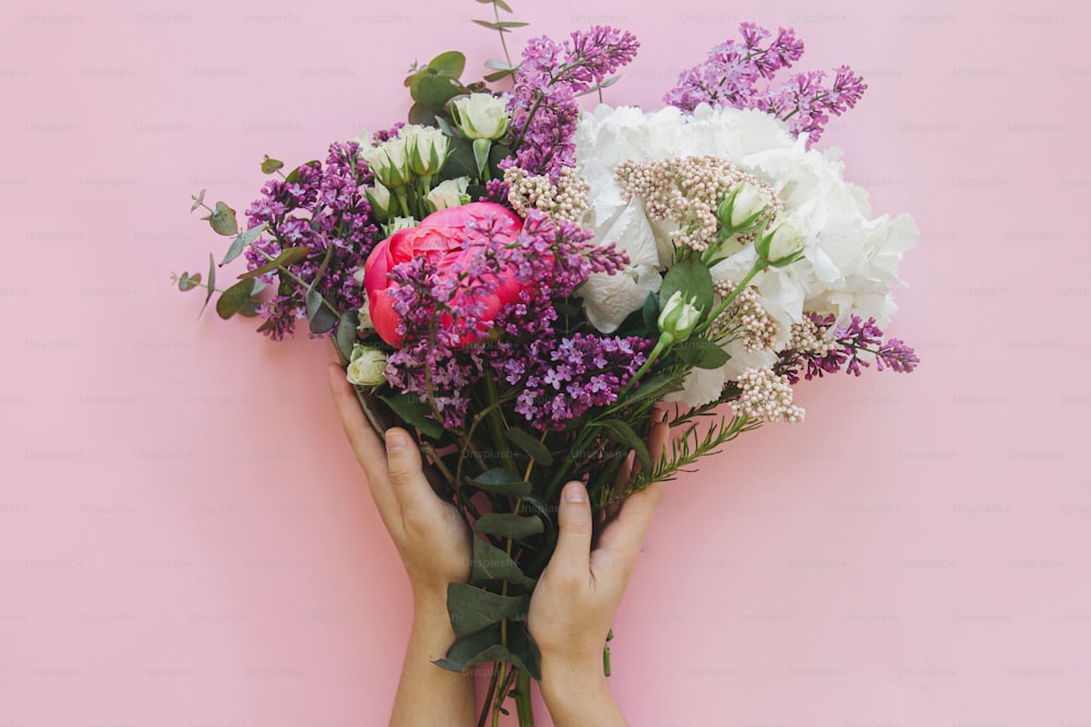 Hände, die modernen frischen Strauß auf leuchtend rosa Hintergrund flach legen. Stilvolle bunte Grußkarte mit Pfingstrose, Flieder, Eukalyptus, Hortensienblüten. Frohen Frauentag. Schönen Muttertag
